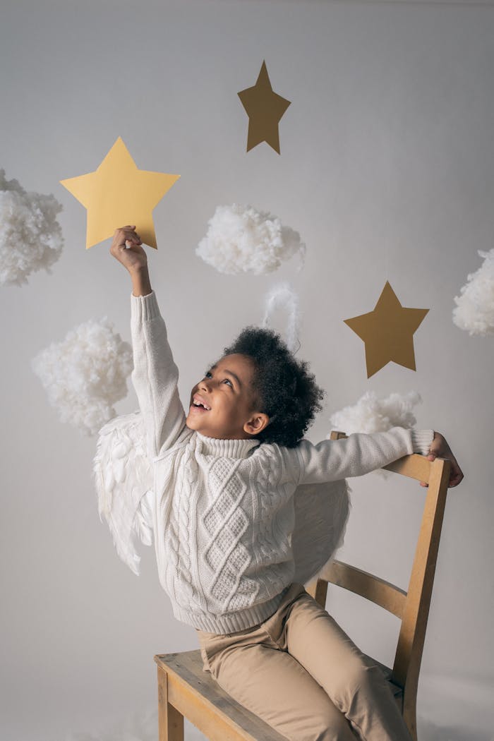 Cheerful African American child with angel wings touching decorative star while looking up from chair on light background